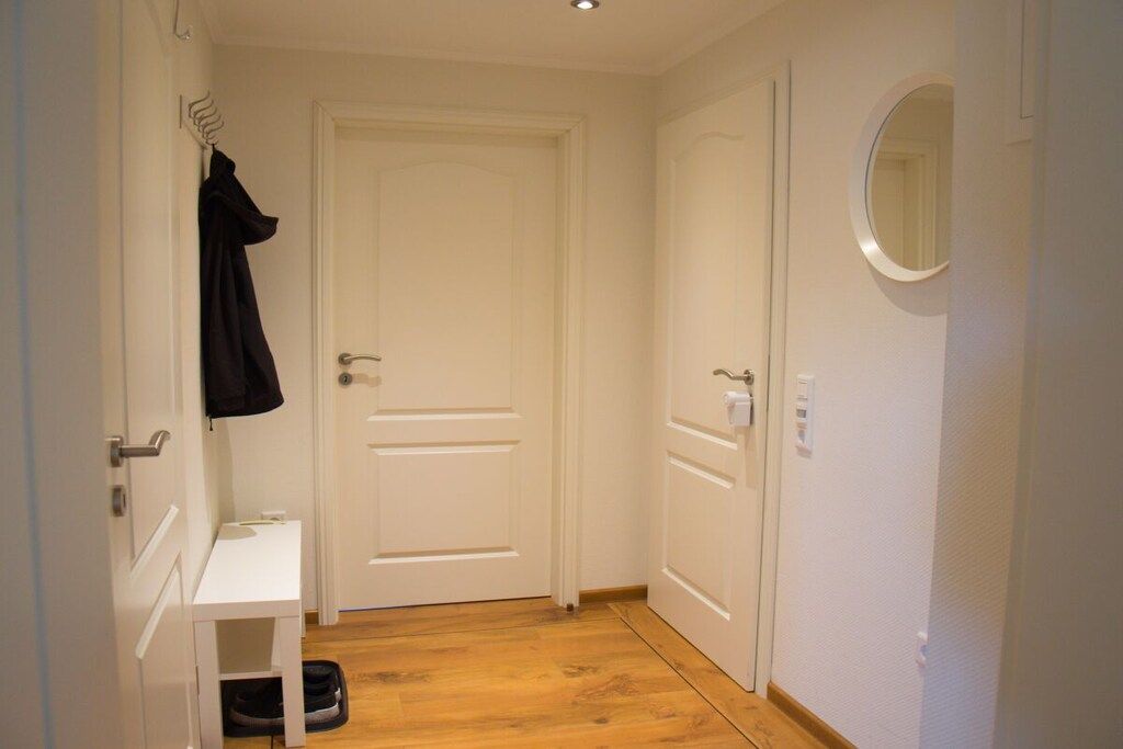 Ein schmaler Flur mit einer weißen Tür, einem kleinen weißen Tisch mit einem schwarzen Gegenstand darauf und einem runden Spiegel an der Wand.