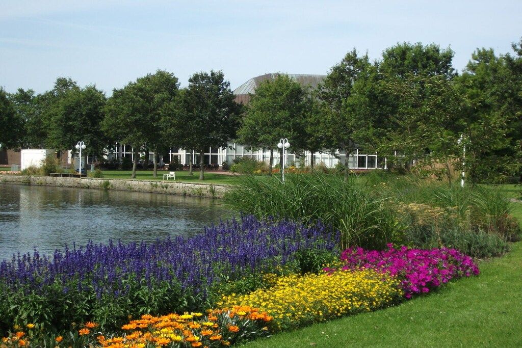 A serene outdoor setting with a pond, vibrant flower beds, and a building with a white fence and lamp posts in the background.
