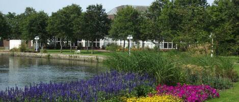 A serene outdoor setting with a pond, vibrant flower beds, and a building with a white fence and lamp posts in the background.