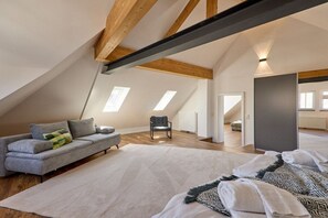 Ein geräumiges Dachzimmer mit schrägem Dach, einem grauen Sofa, einem gemusterten Teppich und einem großen Fenster.