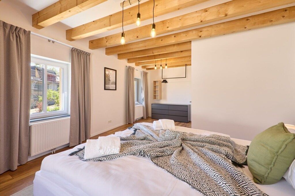 A bedroom with a large bed, wooden beams, a window, a dresser, and a bedside table.