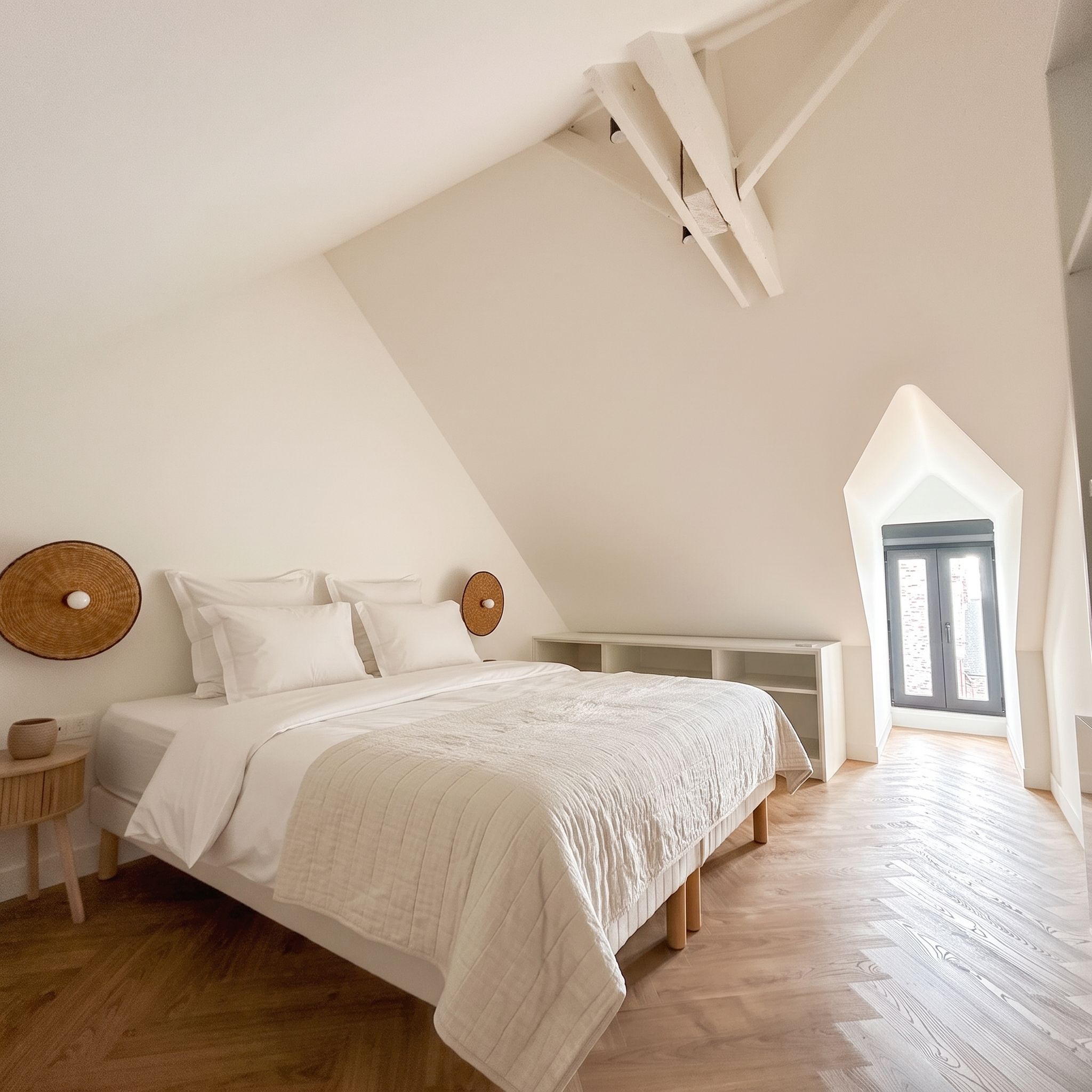 Une chambre avec un plafond en pente, un lit avec une literie blanche, un élément de décoration circulaire sur le mur et une fenêtre donnant sur un paysage.