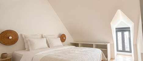 Une chambre avec un plafond en pente, un lit avec une literie blanche, un élément de décoration circulaire sur le mur et une fenêtre donnant sur un paysage.