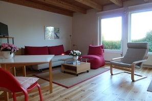 A modern living room with a sofa, red chairs, a glass coffee table, and a view through large windows. - Holiday apartment with 1 bedroom (Fehmarn)