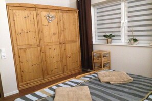 Ein Zimmer mit einem Holzschrank, einem Bett mit gestreifter Bettwäsche, einer kleinen Holzablage und zwei Blumentöpfen auf einem Fensterbrett.