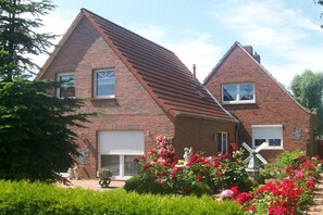A two-story brick house with a visible garage door, a windmill in the garden, and a variety of flowering plants. - Holiday home with 2 bedrooms (Greetsiel)