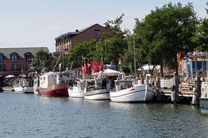 A marina with boats docked, a building with a sloped roof, and trees in the background. - Holiday apartment with 1 bedroom (Heiligenhafen)