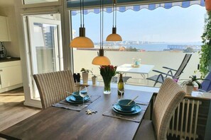 A dining area with a table set for two, a view of the sea, and a balcony with chairs. - Holiday apartment with 1 bedroom (Heiligenhafen)