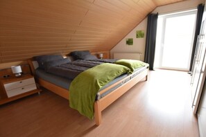 Ein Schlafzimmer mit schrägen Wänden, einem Bett mit grüner Decke, einem Nachttisch und einem Fenster mit Vorhängen.