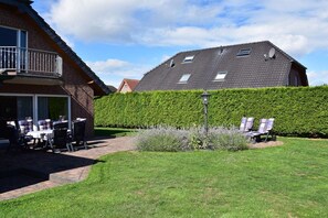 Ein Wohnhaus mit Terrasse, Sitzgelegenheiten im Freien und einem gepflegten Heckenstreifen.