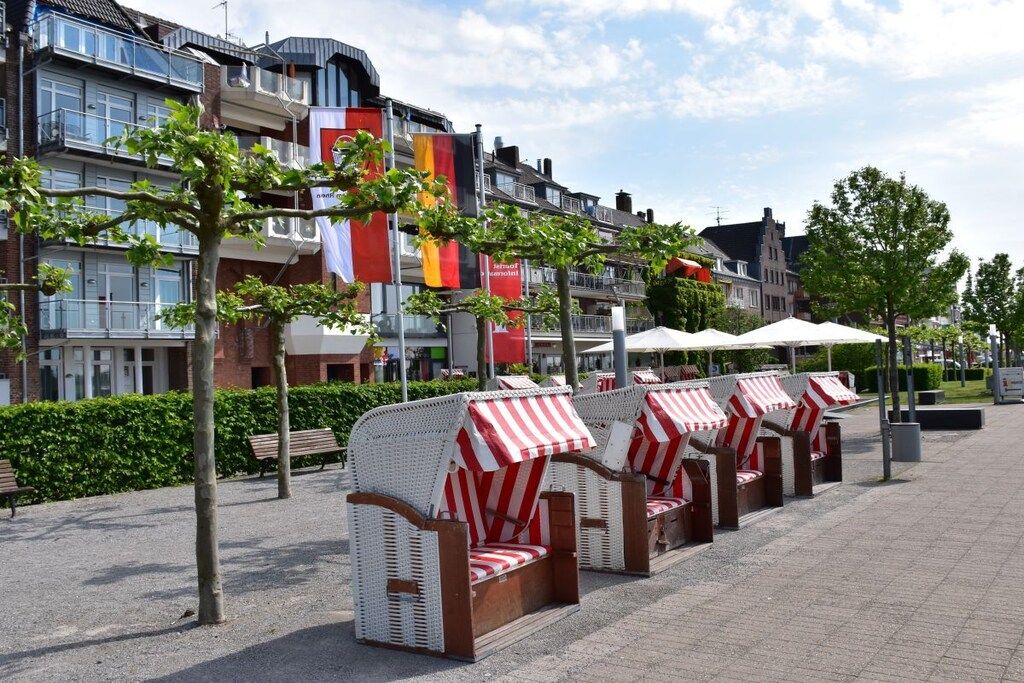 Espace extérieur aménagé avec des chaises en osier blanc et des parasols à rayures rouges et blanches, le tout sur fond de bâtiments ornés de drapeaux.