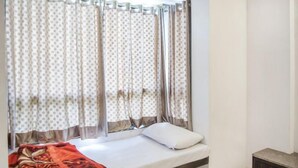 A single bed with a white mattress and a red patterned blanket, a small bedside table, and curtains with a polka dot pattern. - Reveridge Skyinn 6BH Bungalow (Mahabaleshwar)