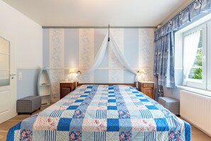 Une chambre à coucher avec un grand lit, deux tables de chevet, une fenêtre avec des rideaux et un mur orné d’un motif floral.
