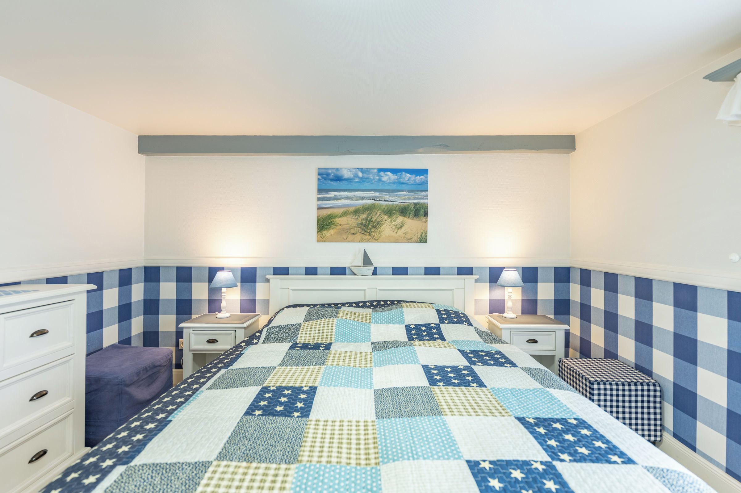 Ein Schlafzimmer mit karierter Wand, einem Bett mit gesteppter Bettdecke mit Sternenmuster, zwei Nachttischen mit Lampen, einem Beistelltisch und einem Fenster mit Blick auf ein Strandgemälde.