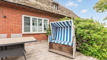 Eine Terrasse mit Tisch und Stühlen, eine Ziegelwand, ein reetgedecktes Dach und ein Strauch mit rosa Blüten.