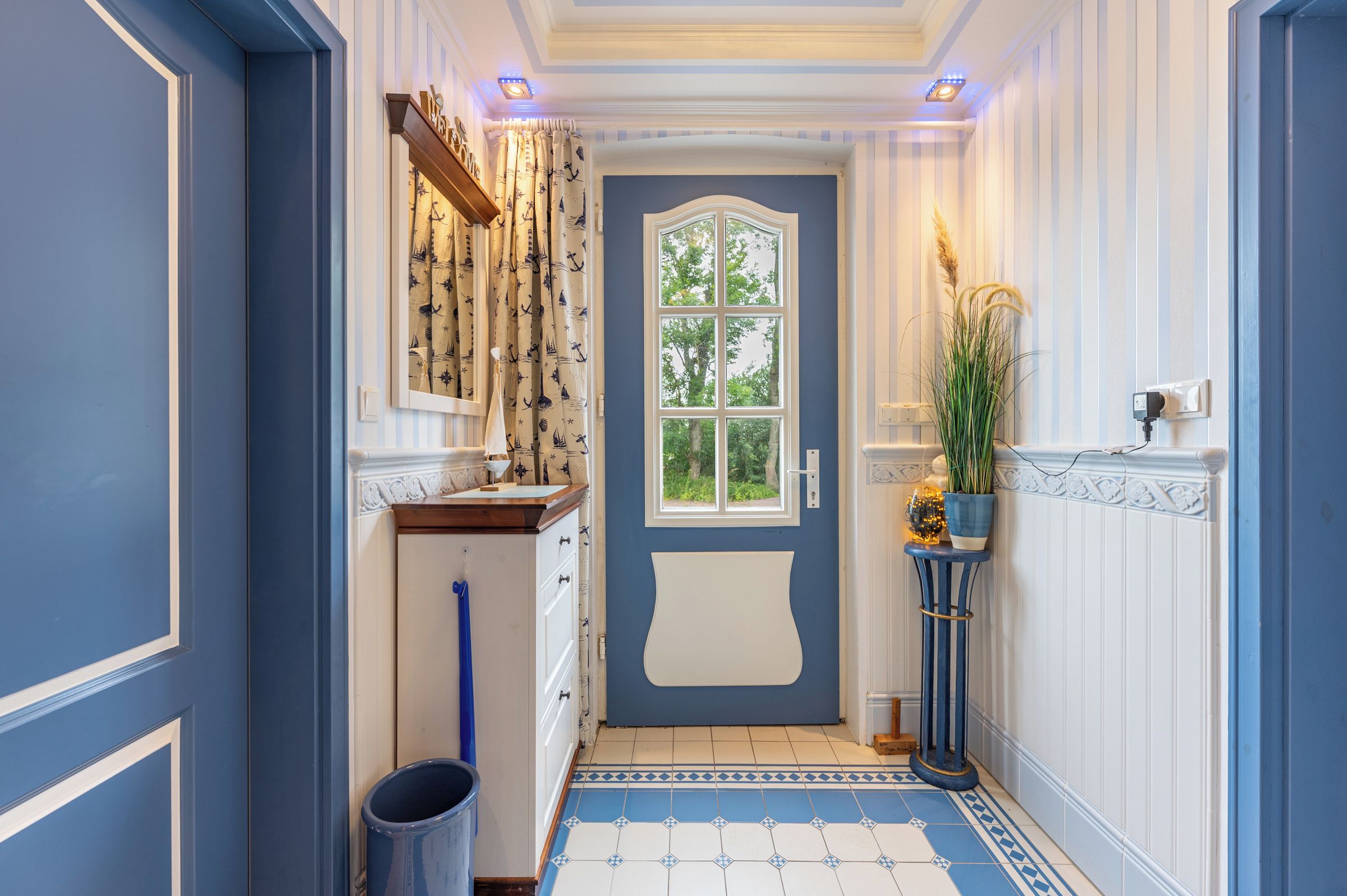 Un couloir avec une porte bleue, des rideaux à motifs, une plante en pot et un sol carrelé.