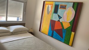 Une chambre à coucher avec un lit, une fenêtre avec des stores et un tableau abstrait aux couleurs vives accroché au mur.