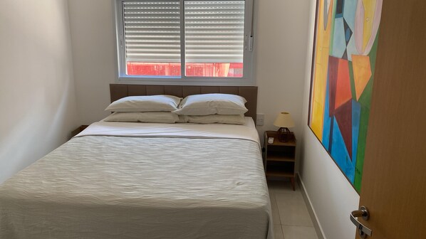 Une chambre à coucher avec un lit, une table de chevet, une lampe, une fenêtre avec des stores et un tableau coloré au mur.