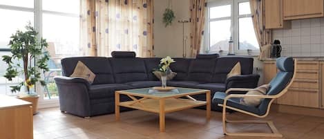 A living room with a sofa, a coffee table, and a chair. There is a window with curtains, a plant, and a view outside.