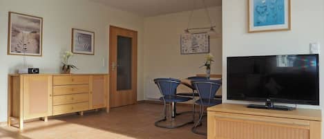 A living room with a television, a wooden cabinet, a dining table with chairs, and artwork on the walls.