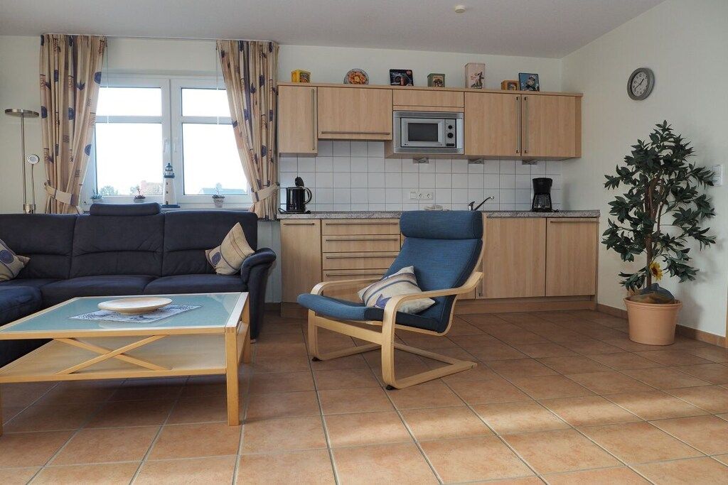 Un salon avec un canapé, une table basse et un fauteuil à bascule. La cuisine est équipée de meubles en bois, d’un four à micro-ondes et d’une plante en pot.