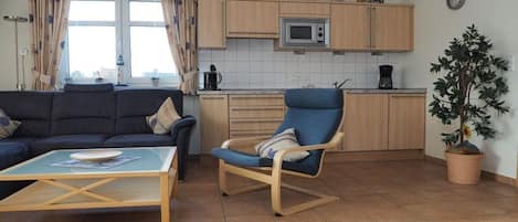 Una sala de estar con un sofá, una mesa de centro y un sillón mecedora. La cocina tiene armarios de madera, un microondas y una planta en maceta.