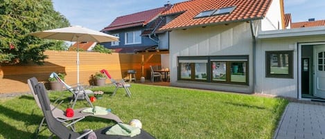 Il s’agit d’une propriété résidentielle dotée d’une pelouse bien entretenue, d’un espace extérieur aménagé avec des chaises et un parasol, ainsi que d’une maison avec un toit recouvert de tuiles rouges et de grandes fenêtres.