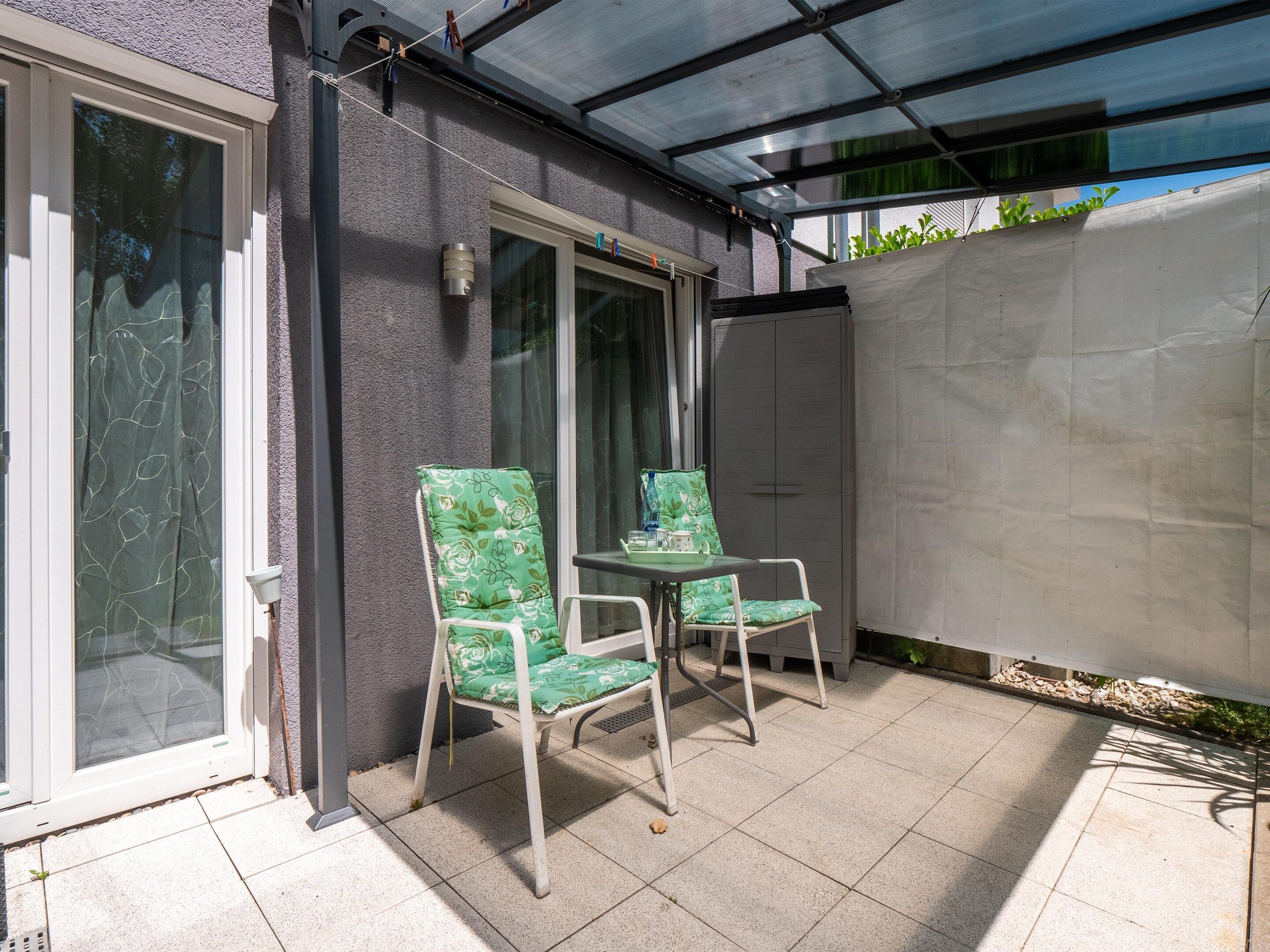 Ein überdachter Patio-Bereich mit zwei Stühlen, einem Tisch, einer Glastür und einer weißen Wand.