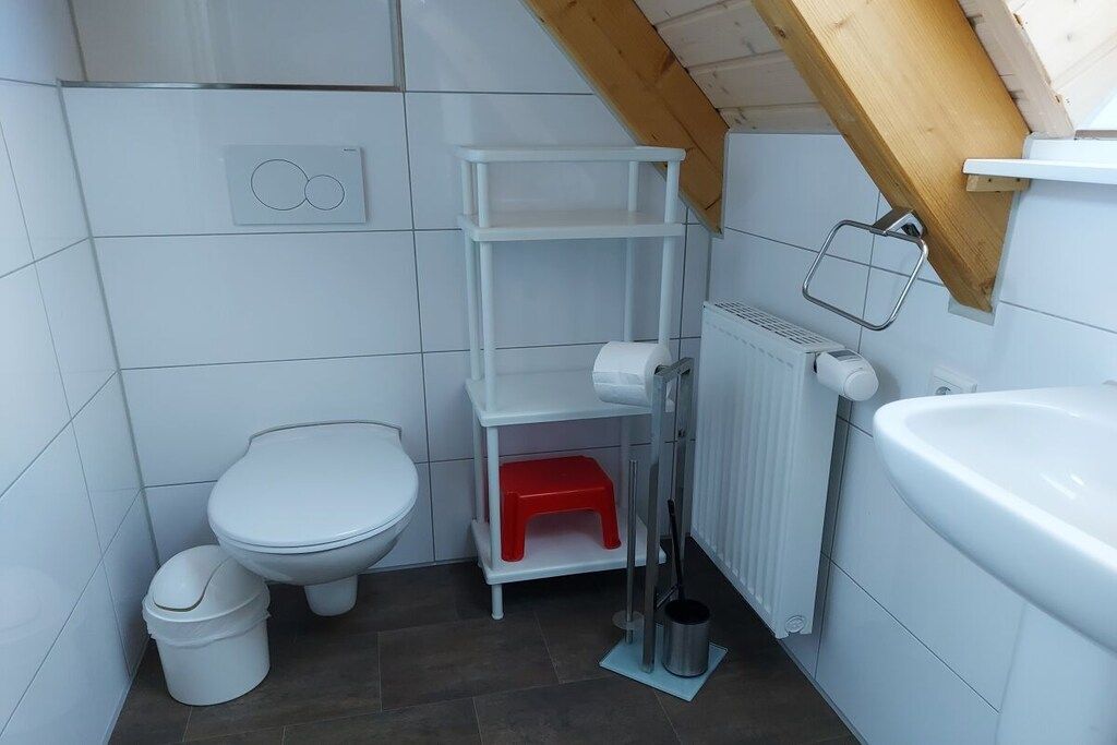 Ein kleines, modernes Badezimmer mit einer weißen Toilette, einem weißen Waschbecken und einem weißen Heizkörper. Auf dem Boden steht ein roter Tritt und ein weißer Eimer.