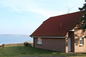 A single-story brick building with a red-tiled roof, a small window, and a door, situated on a grassy area with a body of water in the background. - "Desire for a house by the lake" (Sternberg)