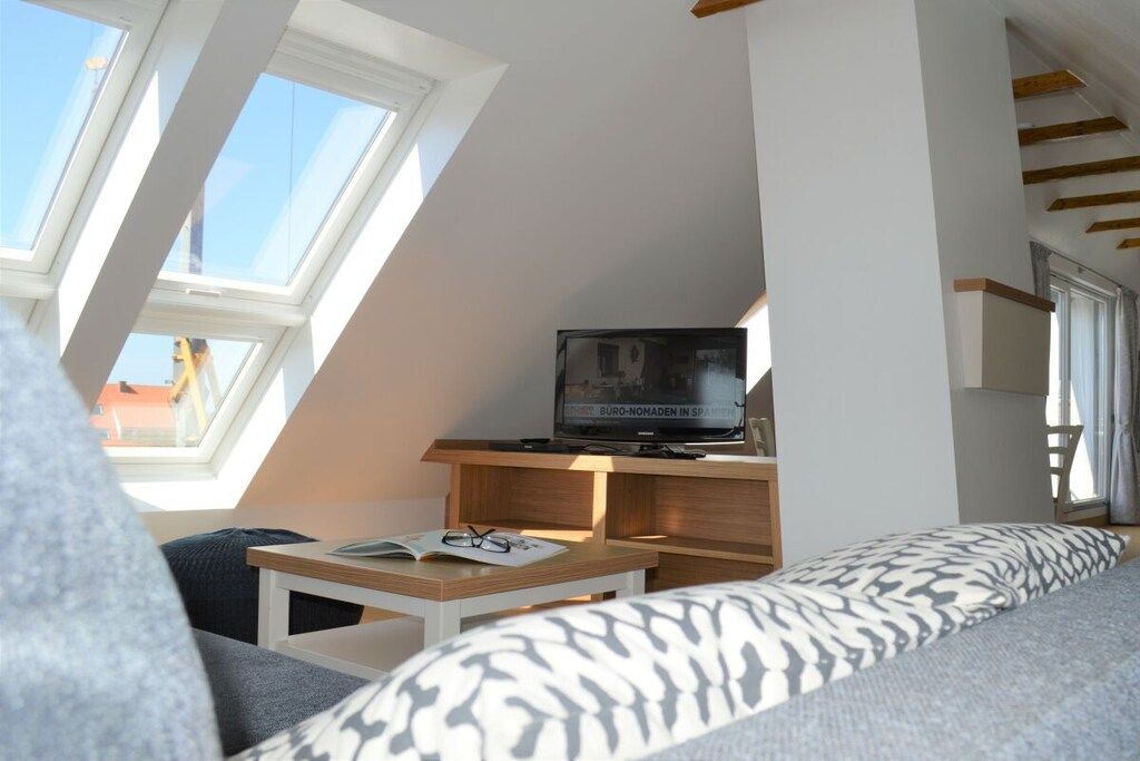 Una habitación con techo inclinado, un televisor sobre un mueble de madera, un sofá con cojines estampados y una mesita con un libro y gafas.