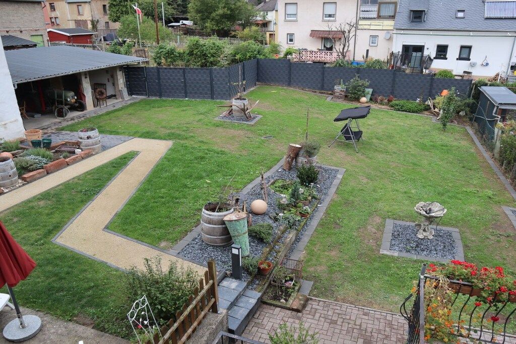 Een achtertuin van een woning met een verhard terras, een klein plantenveld met beplanting en een vogelbad, en een zichtbare garage.