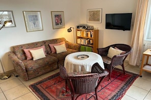 A living room with a sofa, two chairs, a small round table, a bookshelf, and a TV mounted on the wall. - Holiday apartment in Juist with swimming pool (Juist)