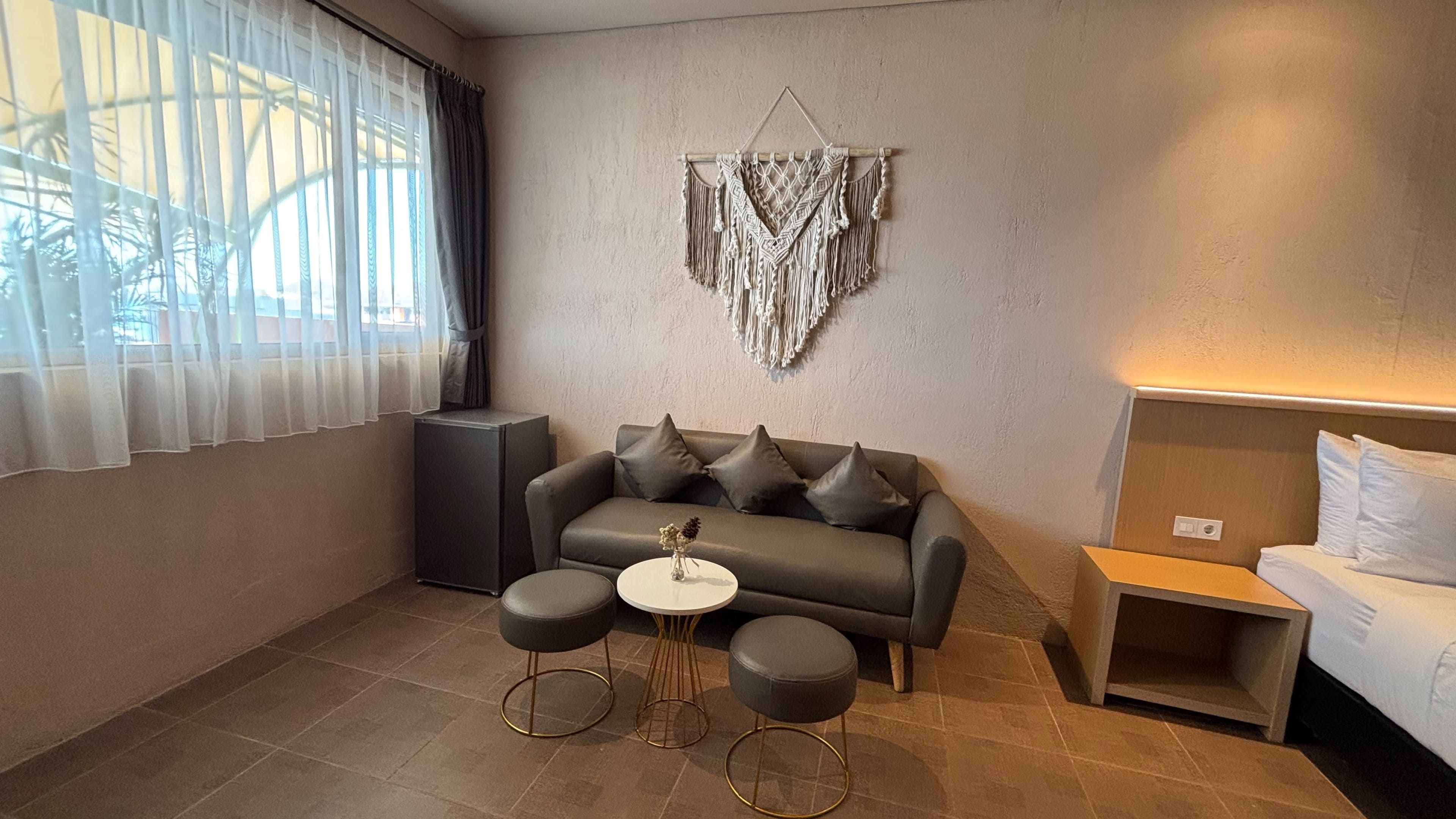 Una habitación moderna con un sofá gris, dos otomanas negras redondas, una mesita blanca y un colgador de macramé en la pared.