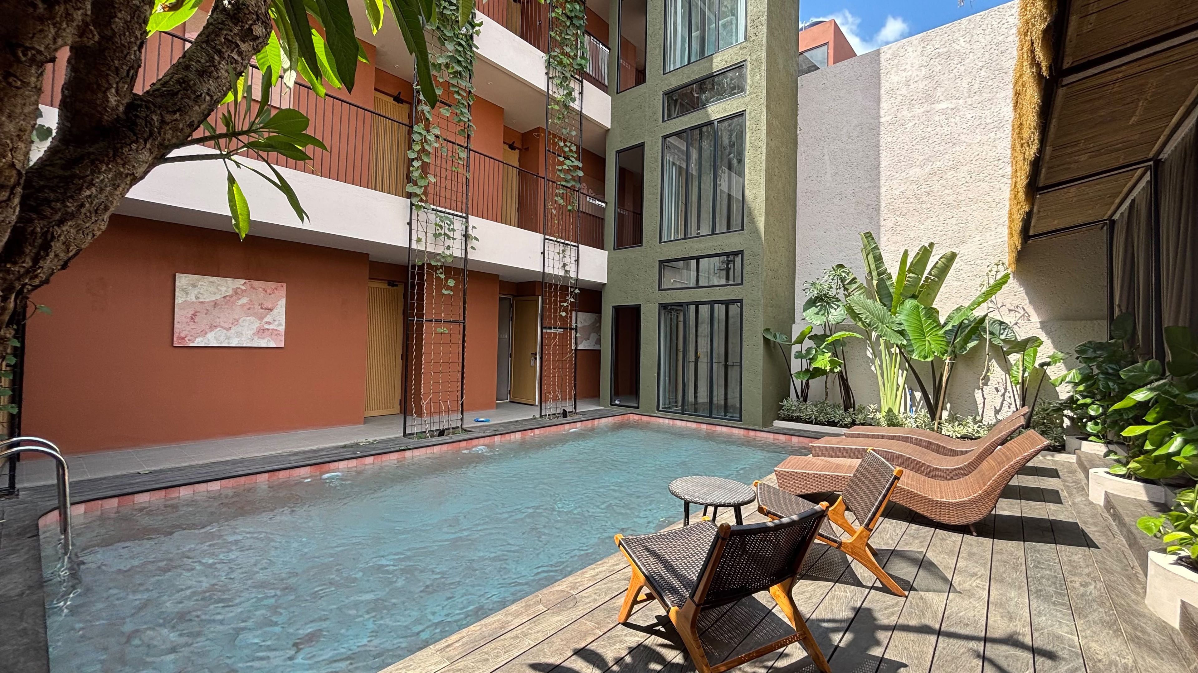 A swimming pool with lounge chairs and tables, surrounded by a building with balconies and large windows.