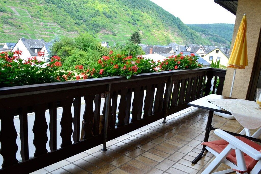 A balcony with a view of a hillside, a table with a chair, and a yellow umbrella.