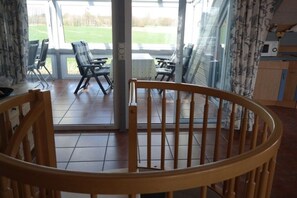 Ein Zimmer mit einem Holzstuhl im Vordergrund, einem Essbereich mit Stühlen und einem Tisch und Blick auf ein grünes Feld durch große Fenster.