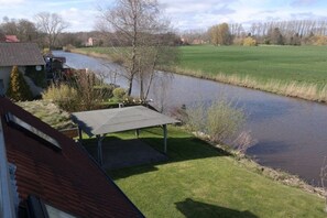 Ein Haus am Flussufer mit überdachter Terrasse, einem roten Dach und einer grünen Wiese.