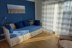 A room with a blue sofa, a painting on the wall, and a window with striped curtains. - Holiday apartment with 1 bedroom (Carolinensiel)