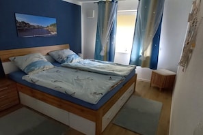 A bedroom with a bed, a nightstand, a small wooden stool, and a window with curtains. - Holiday apartment with 1 bedroom (Carolinensiel)