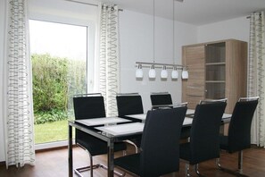 A modern dining area with a table and chairs, a wooden cabinet, and a view of greenery outside. - Holiday home in Cuxhaven with swimming pool (Cuxhaven)