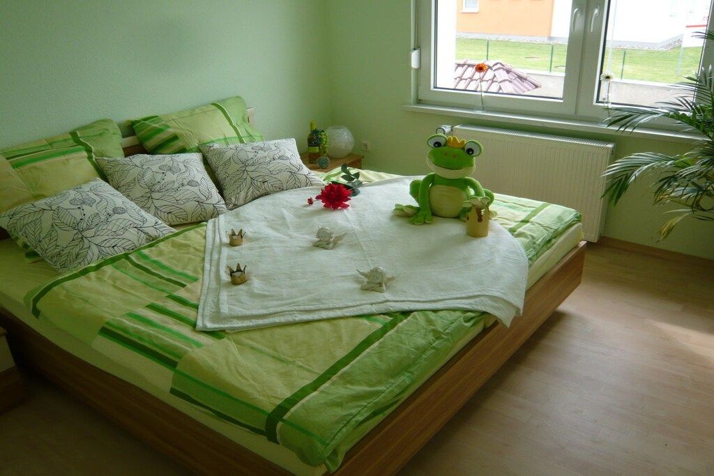 Ein Bett mit einer grün-weißen gestreiften Tagesdecke, einem grünen Plüschfrosch und einer weißen Decke mit roter Blume.