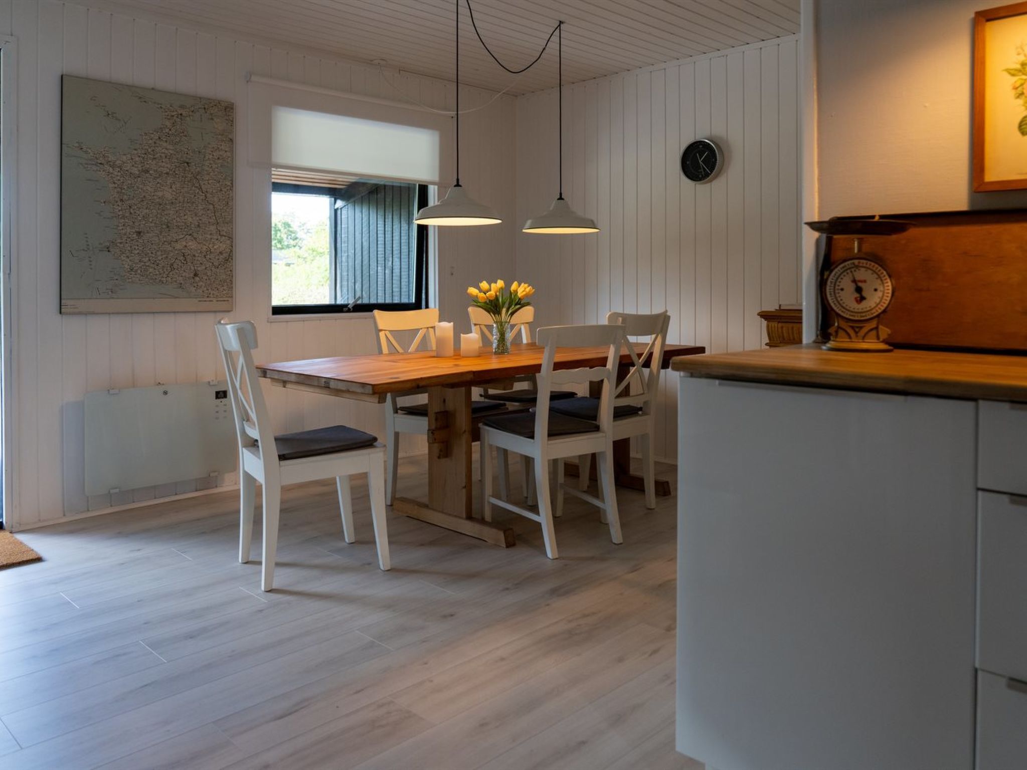 Een eetgedeelte met een houten tafel, witte stoelen, een raam met zonwering, hanglampen en een wandklok.