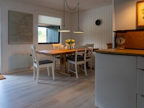Un comedor con una mesa de madera y sillas blancas, una ventana con persianas, lámparas colgantes y un reloj de pared.