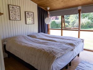 Una cama doble con una colcha acolchada en una habitación con suelo de madera, una ventana con vistas a zonas verdes y dos cuadros con motivos florales en la pared.
