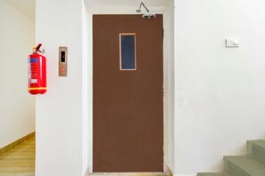 A brown door with a small window, a fire extinguisher mounted on the wall to the left, and a light switch to the right. - Townhouse Oak LB Nagar Metro Station (Hyderabad)
