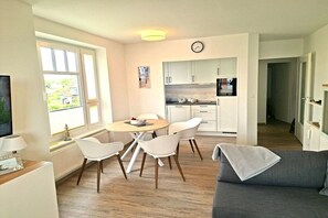 A modern living room with a grey sofa, a dining area with a round table and white chairs, and a kitchen with white cabinets and a clock on the wall. - Holiday apartment with 1 bedroom (Wurster Nordseeküste)