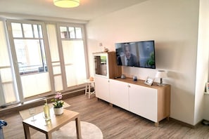 A modern living room with a flat-screen TV, wooden furniture, and a view through large windows. - Holiday apartment with 1 bedroom (Wurster Nordseeküste)