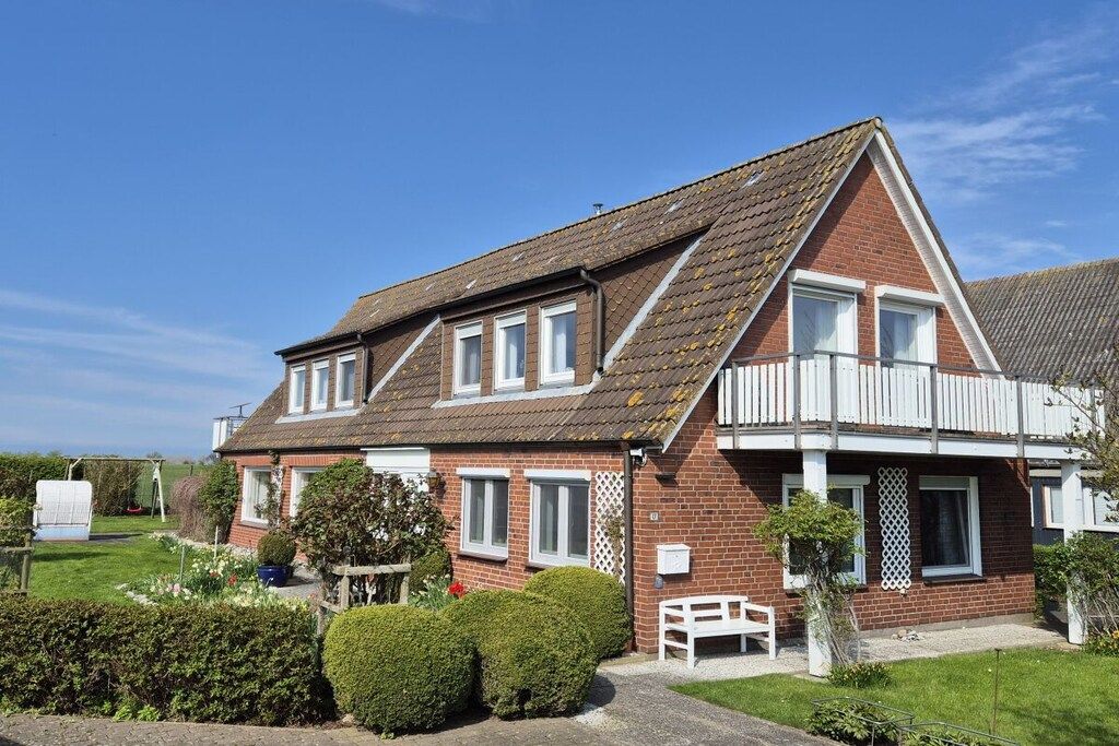 A two-story house with a sloped roof, a balcony on the upper floor, and a small garden area with a bench.
