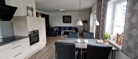A modern kitchen with a dining area, featuring a built-in oven, a flat-screen TV, and a dining table with chairs.